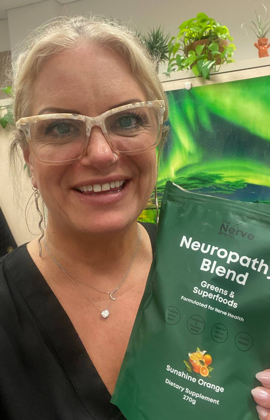 A smiling blonde woman with glasses holding a green pouch of Neuropathy Blend dietary supplement.