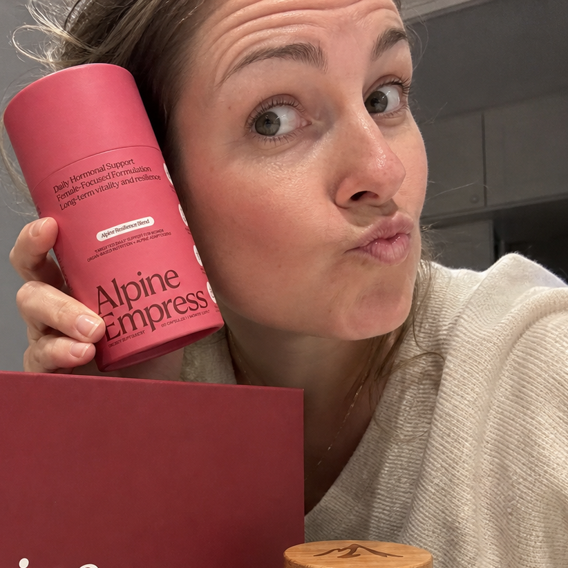 A woman makes a playful face at the camera while holding a pink container of Alpine Empress supplement.