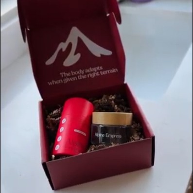 A red container and a glass jar labeled 'Alpine Empress' sit inside an open burgundy gift box.