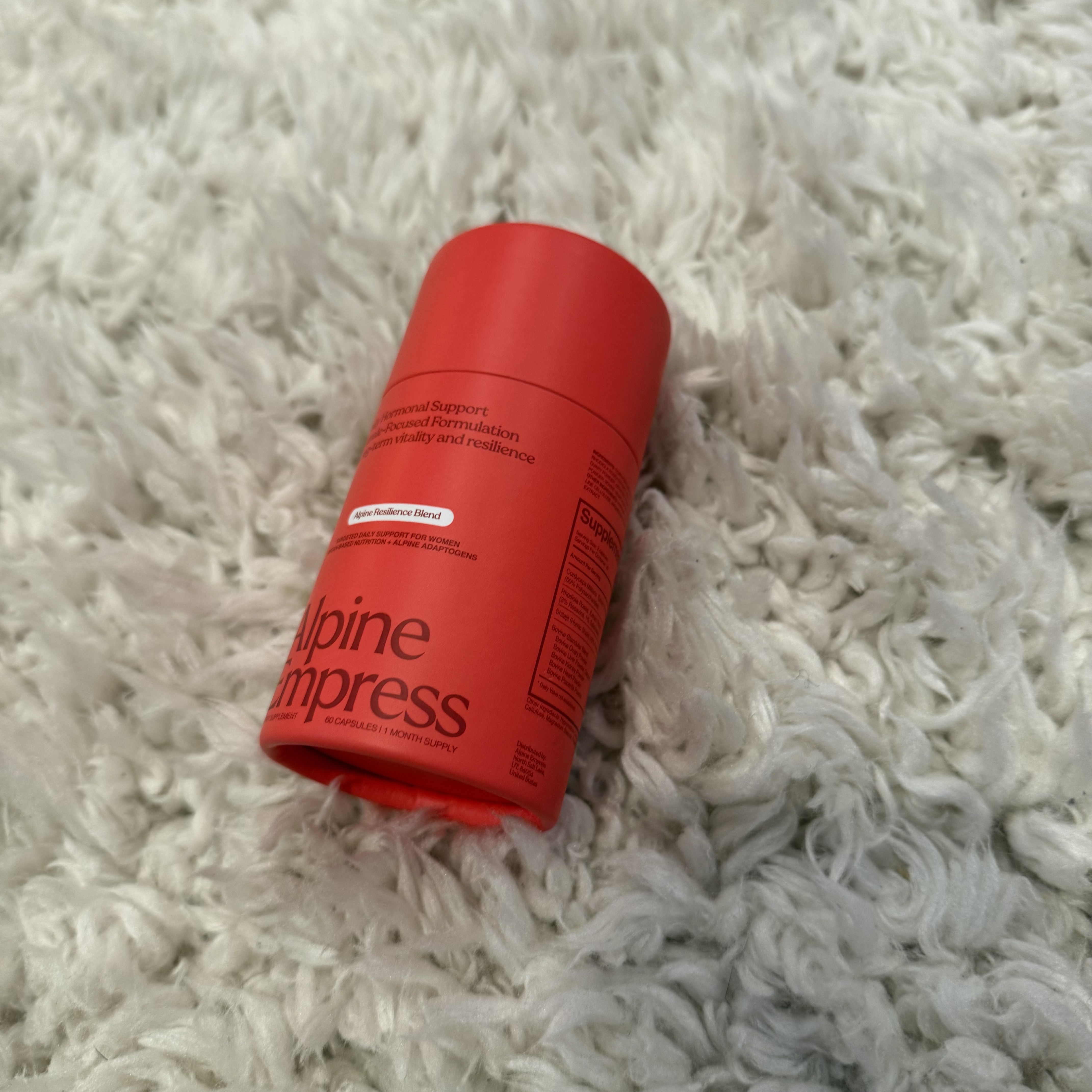 A red bottle of Alpine Empress supplements lying on a fluffy white rug.