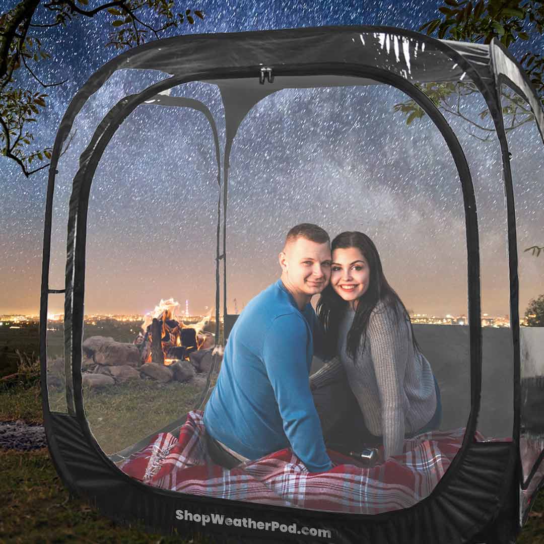 A couple smiles from inside a clear pop-up tent at night under a starry sky.