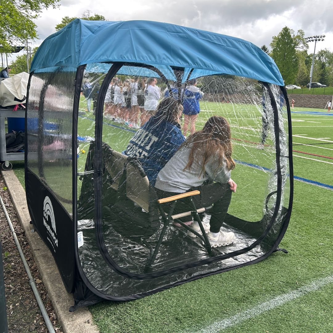 Two people sit inside a personal, clear-sided weather tent on the sideline of a sports field.