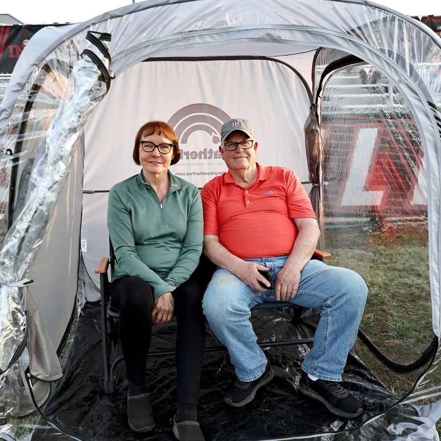 An older couple sits in chairs inside a personal, clear-fronted weather pod at an outdoor event.