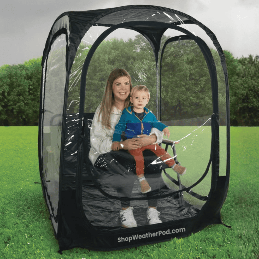 A woman holding a child sits inside a black-framed, clear plastic weather pod on a grassy field.