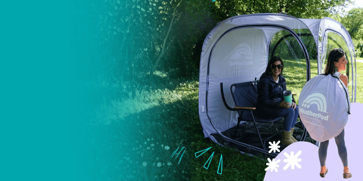 A woman sits in a chair inside a clear, pop-up personal tent on a grassy field.