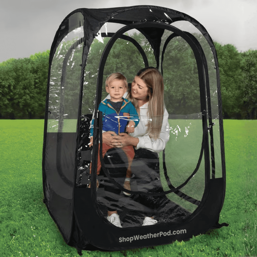 A woman holds a young child while sitting inside a clear, personal pop-up weather tent on a lawn.