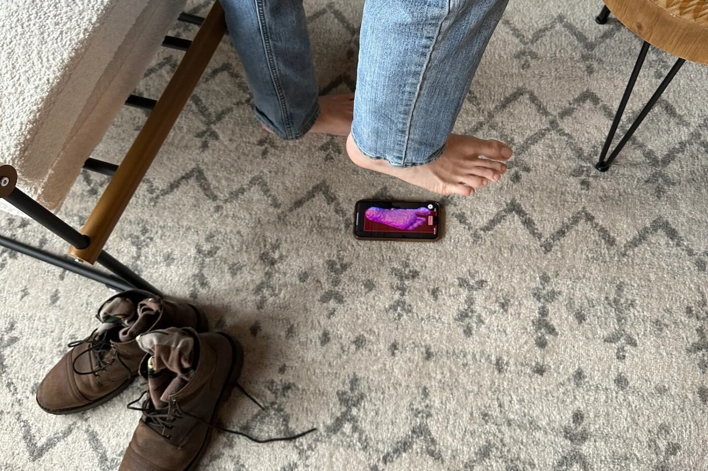 A person steps on a smartphone displaying a purple image, next to boots on a patterned carpet.