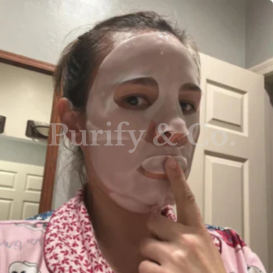 Person wearing a facial sheet mask, touching their lips, in a bathroom.