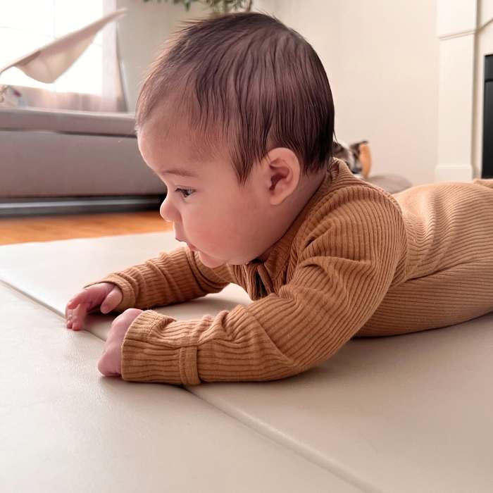 A baby in a brown ribbed outfit lies on its stomach on a light-colored mat indoors.