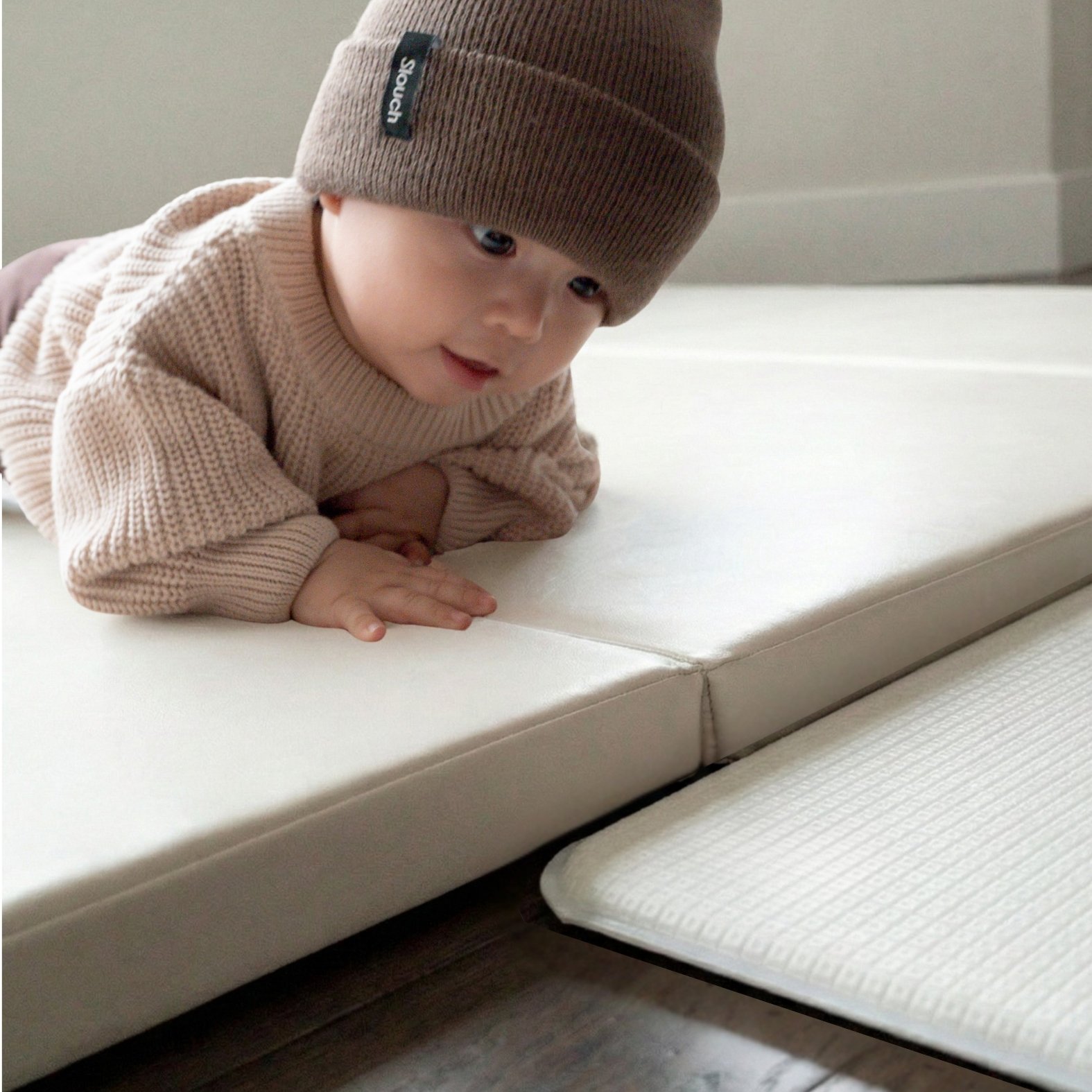 A baby wearing a brown beanie and a cream sweater lies on their stomach on a white play mat.