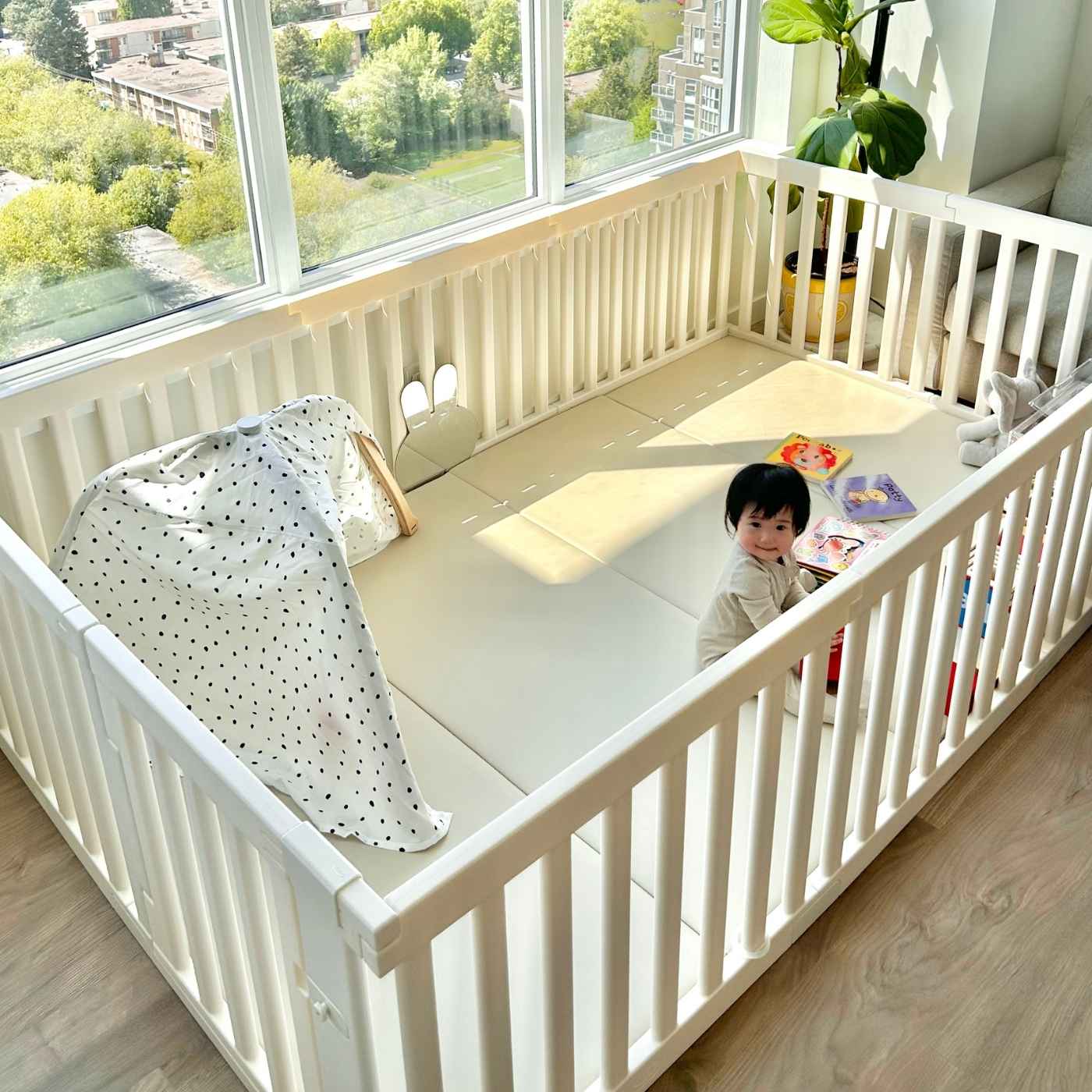 A baby sits inside a large white playpen with toys next to a large window.