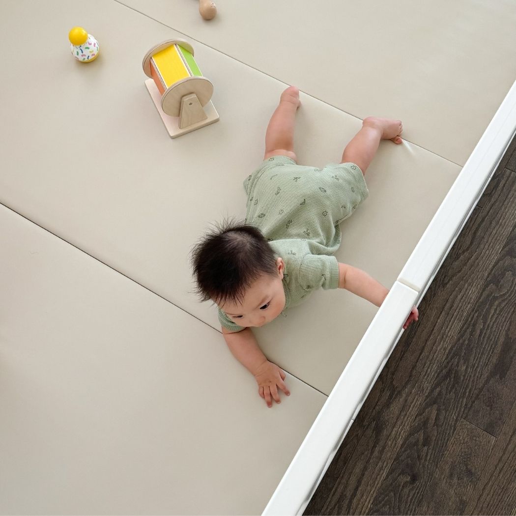 An overhead view of a baby in a green outfit lying on a light-colored play mat.