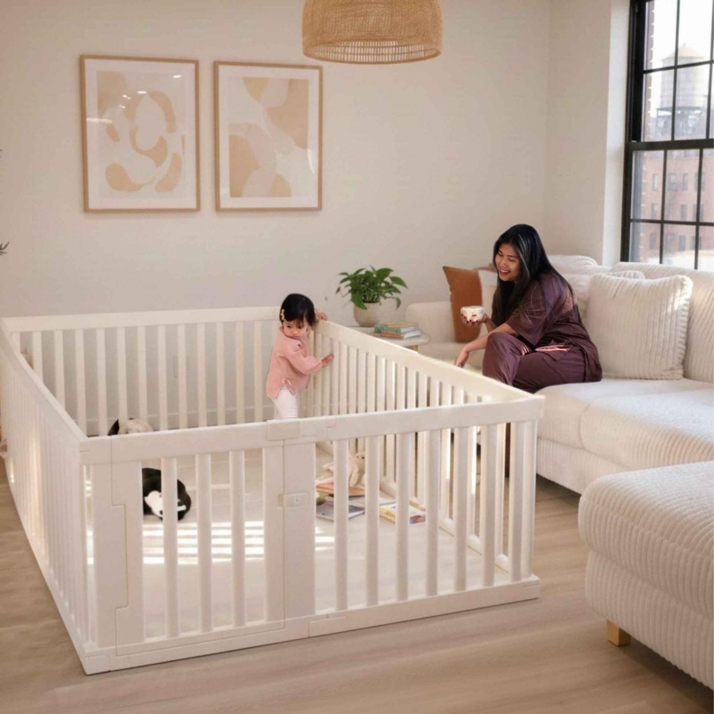 A woman on a couch smiles at a toddler standing in a large white playpen in a living room.