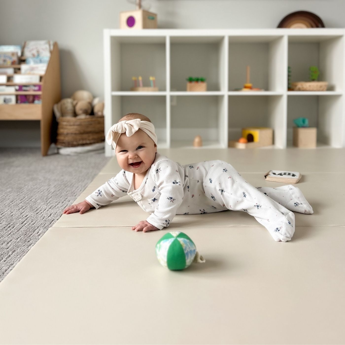 A smiling baby wearing a bow headband lies on a beige play mat in a playroom.