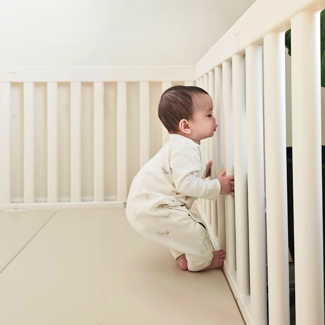 A baby in a white onesie squats in a white crib, holding onto the bars.
