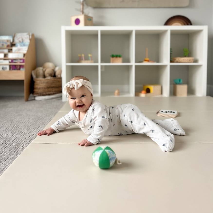 A smiling baby in a floral onesie and a headband lies on a playmat in a playroom.