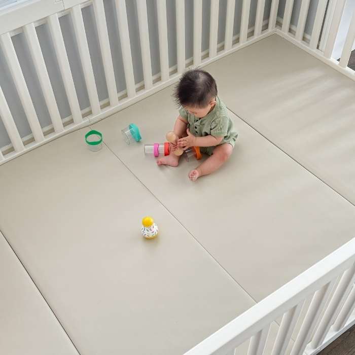 A baby sits on a padded floor mat inside a white playpen, surrounded by toys.