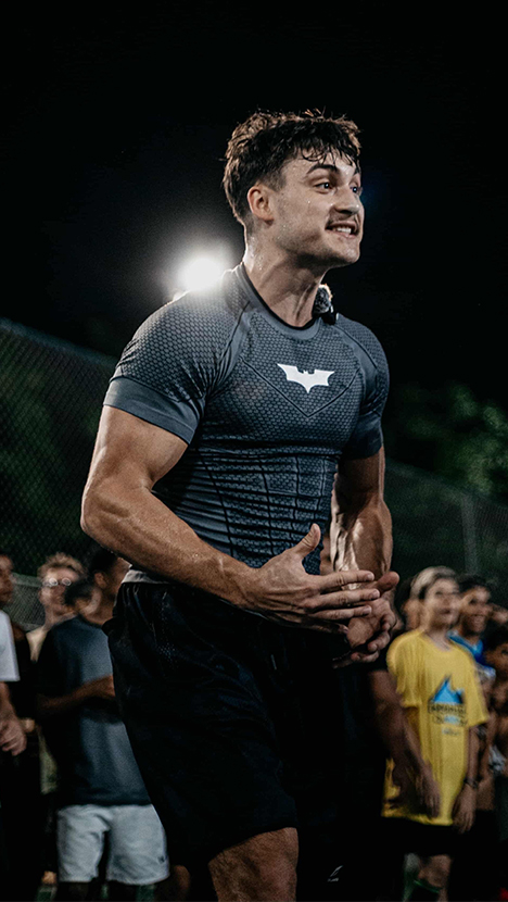 A muscular man in a gray Batman shirt with an intense expression, standing outdoors at night.