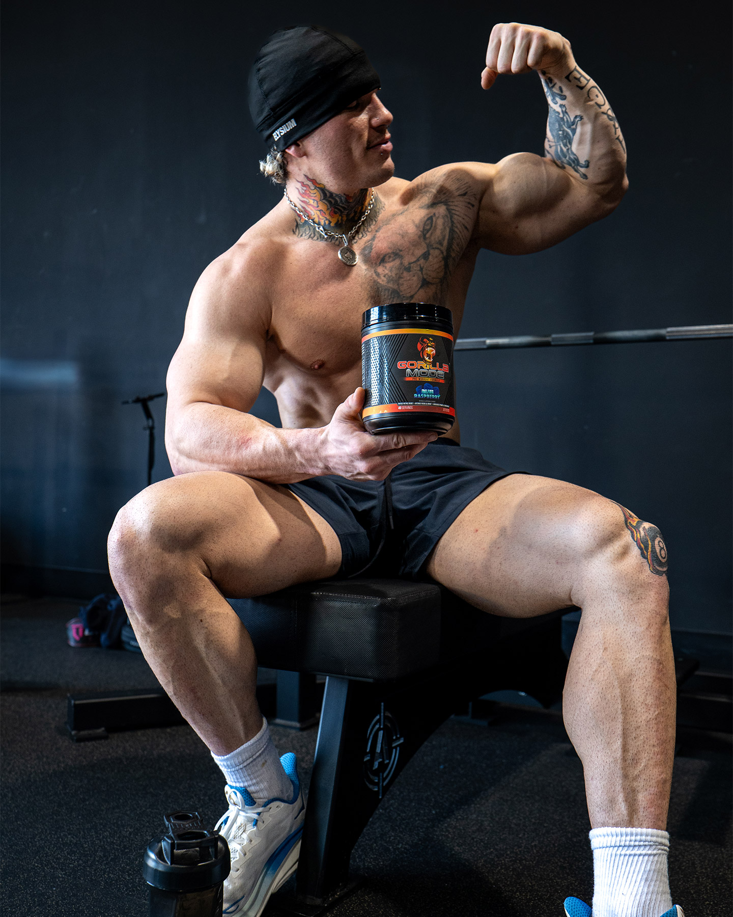 Bodybuilder flexing arm while holding a supplement container, sitting on a gym bench.