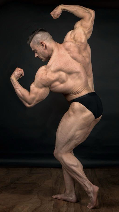 A male bodybuilder in a side pose, flexing his biceps and back muscles against a dark background.