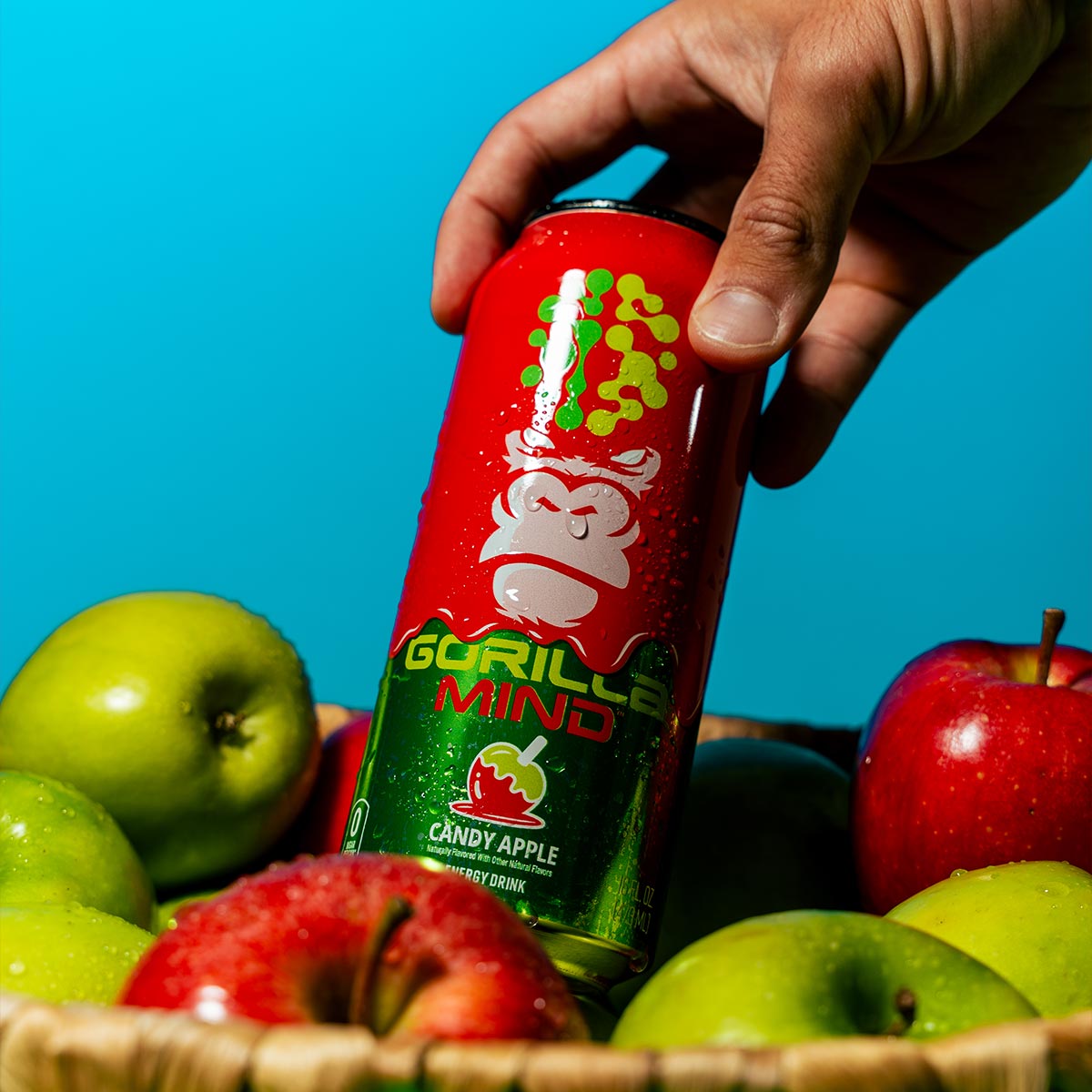 A hand picks a can of Gorilla Mind Candy Apple energy drink from a basket of fresh apples.