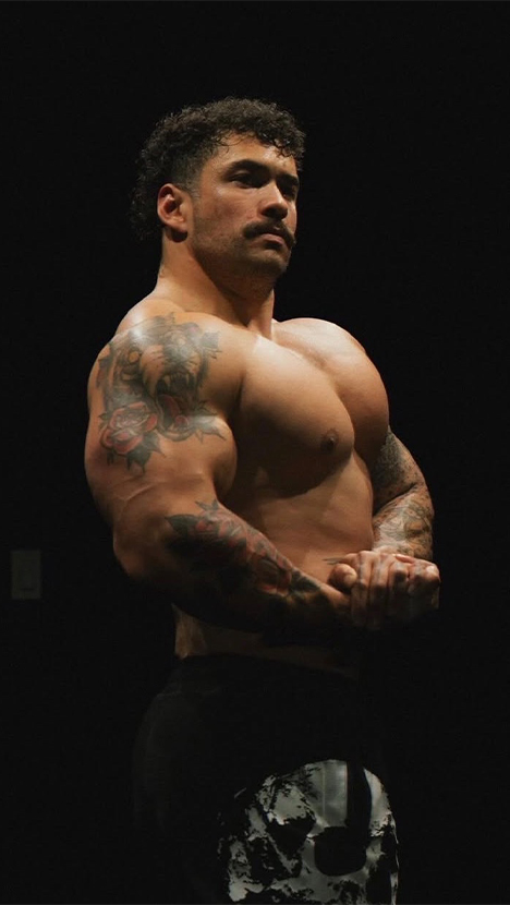 A muscular man with tattoos and a mustache flexes his arms against a black background.