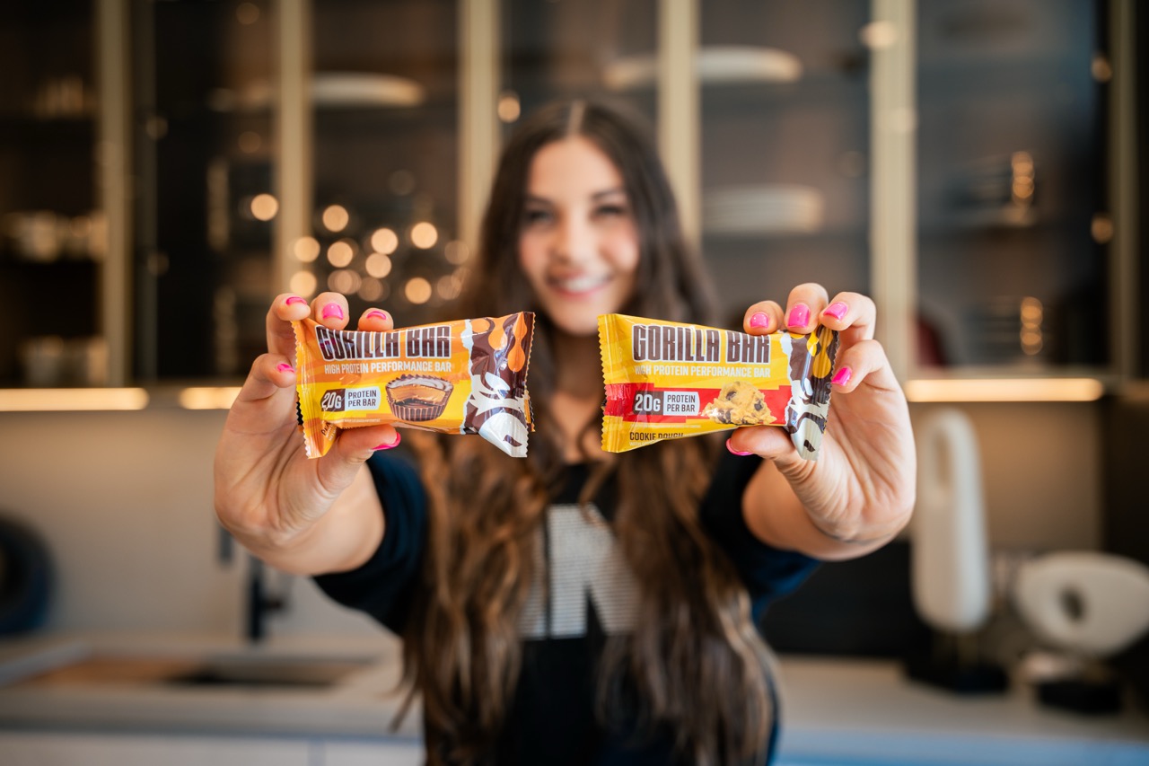 A smiling woman holds two 'Gorilla Bar' high protein performance bars towards the camera.