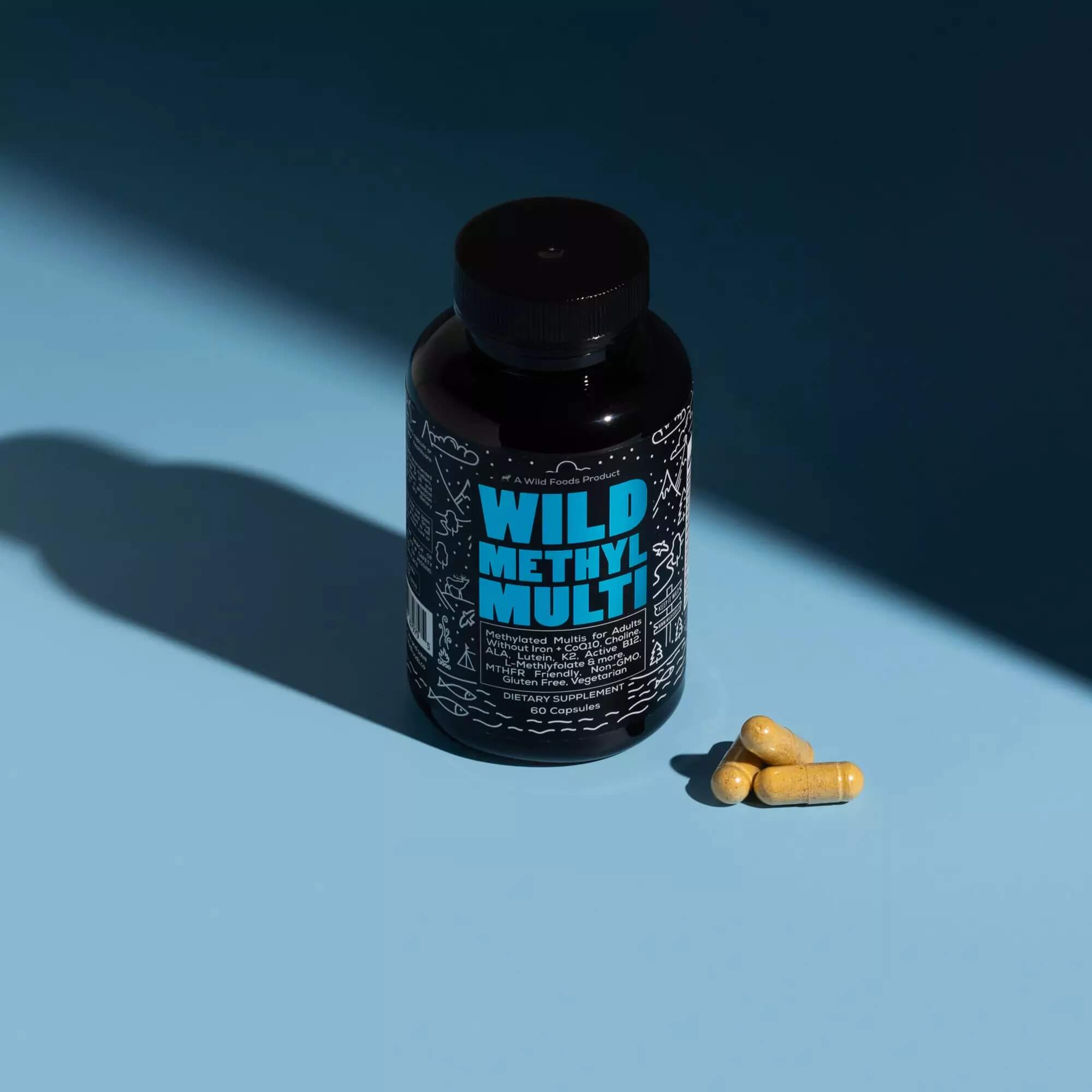 Black bottle labeled 'Wild Methyl Multi' with three yellow capsules on a blue surface.