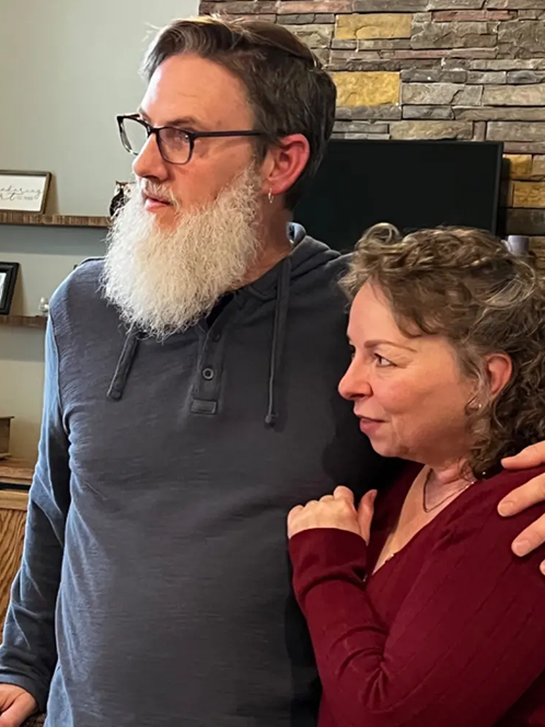 A man with a white beard and a woman standing together indoors.