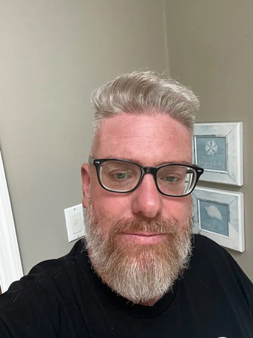 A person with glasses and a beard, standing indoors.