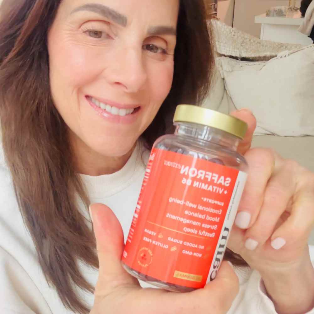A smiling woman in a white sweatshirt holds up a bottle of supplements with a red label.