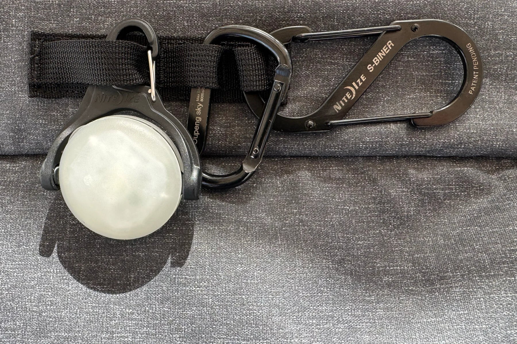 Carabiner and round light attached to a black strap on gray fabric.