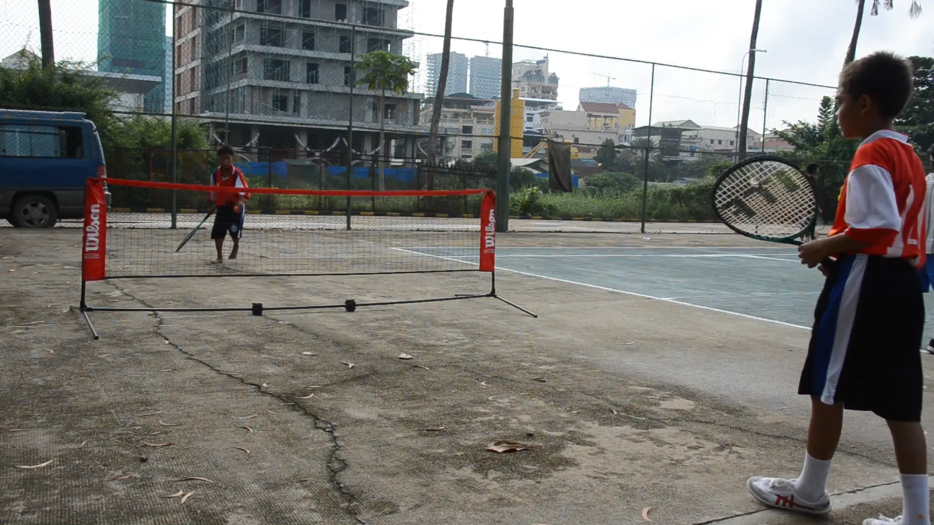 Two children playing tennis on an outdoor court near a construction site.