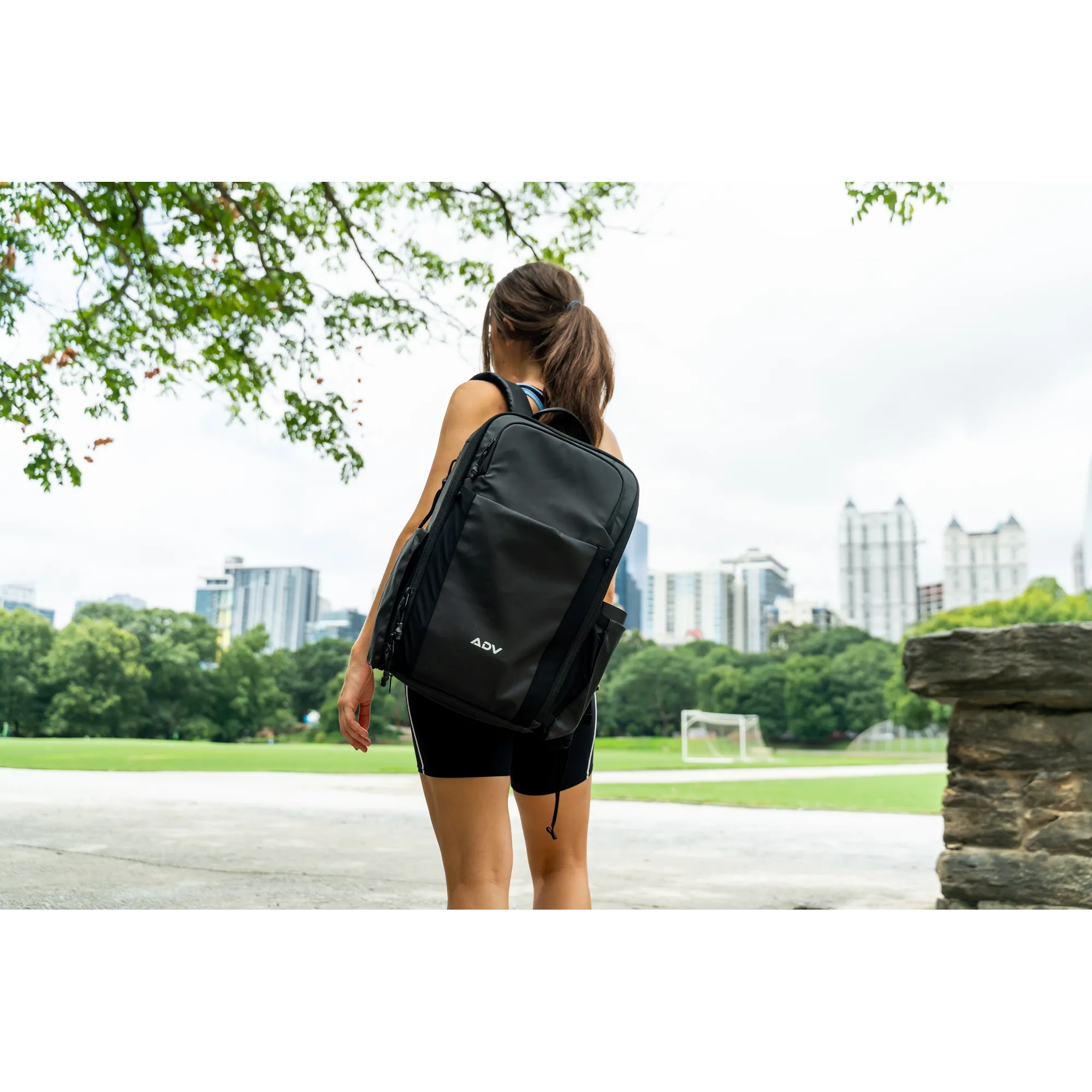 Person with a large backpack standing in a park, city skyline in the background.