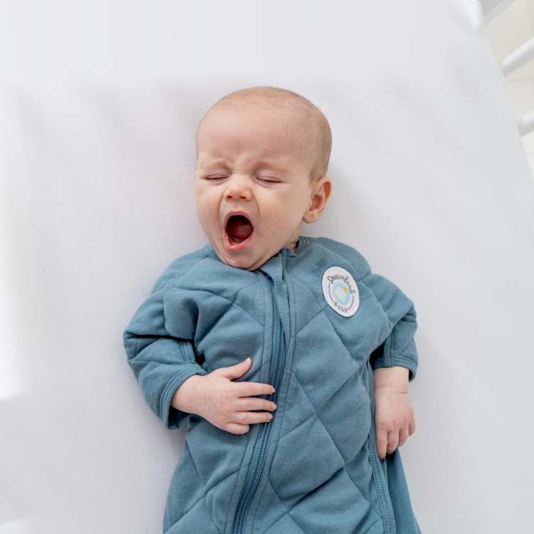 Baby in blue outfit yawning on a white surface.