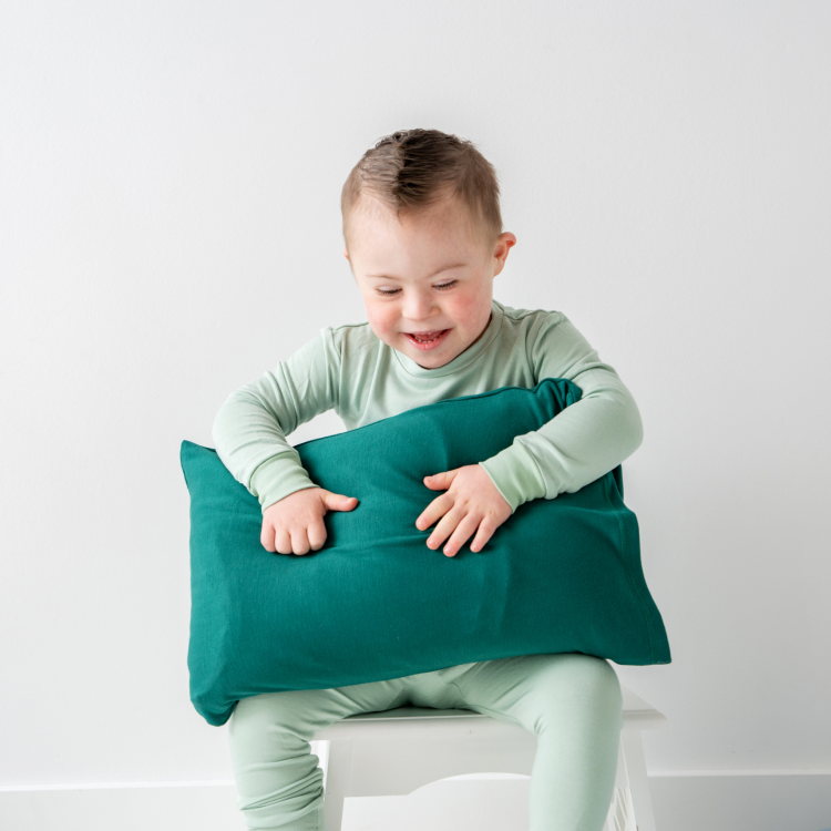 Child in light green pajamas holding a teal pillow, sitting on a white stool.