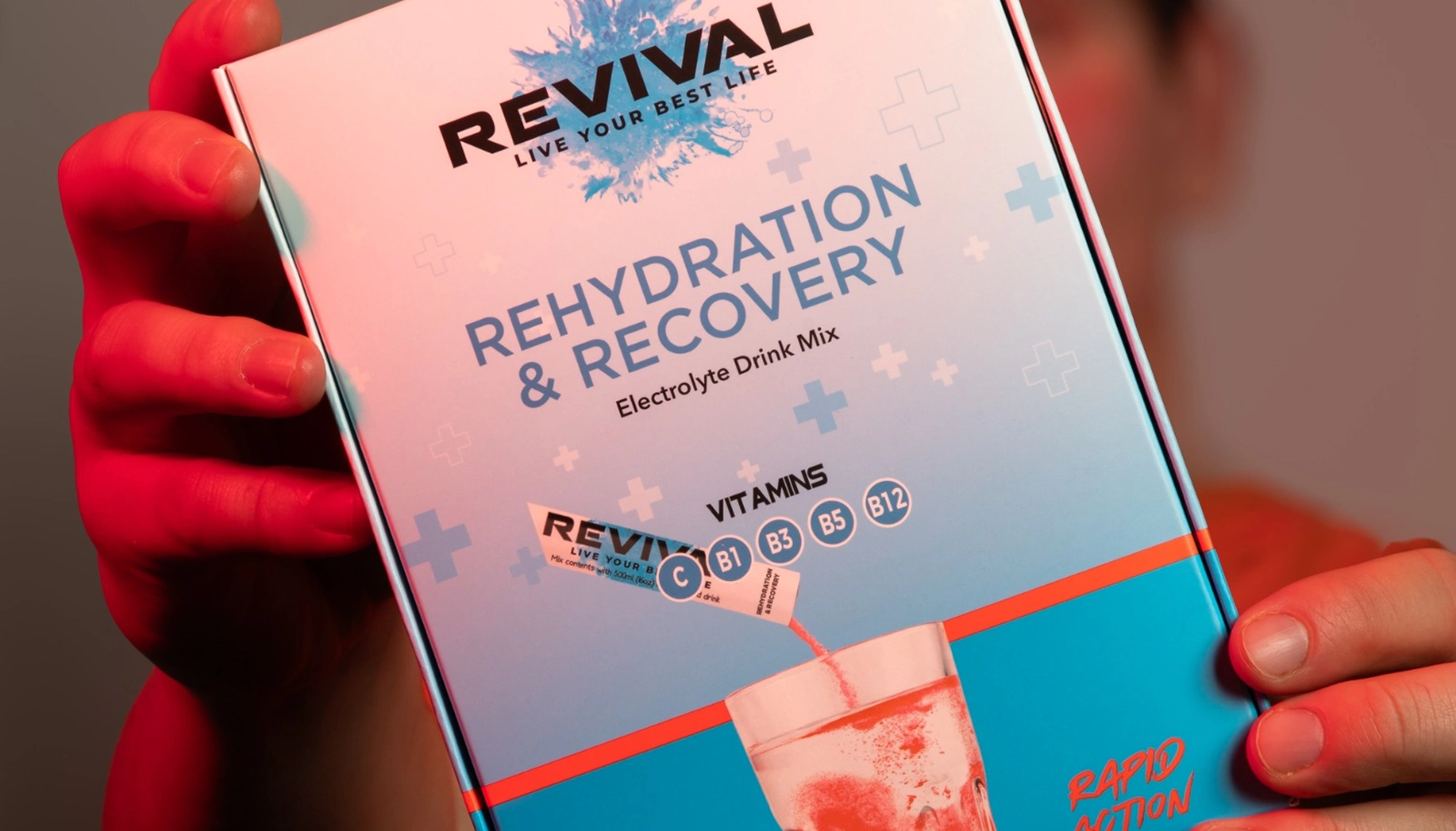 A person holds a box of Revival Rehydration & Recovery electrolyte drink mix under red and blue lighting.