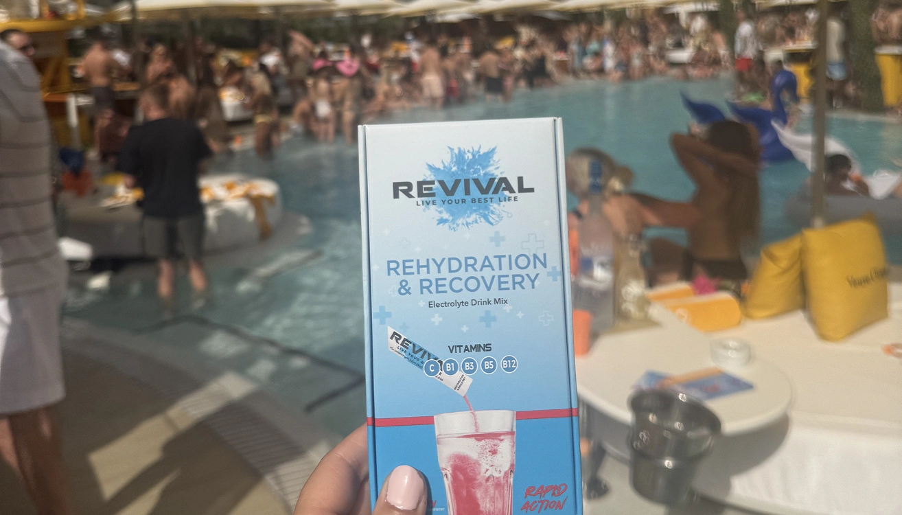 A hand holds a box of Revival electrolyte mix in front of a crowded pool party.