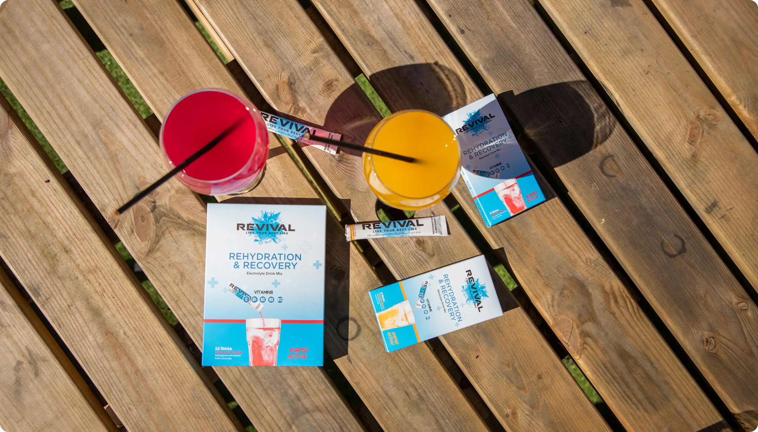 An overhead shot of Revival Rehydration & Recovery drink mix products next to two colorful drinks on a wooden table.