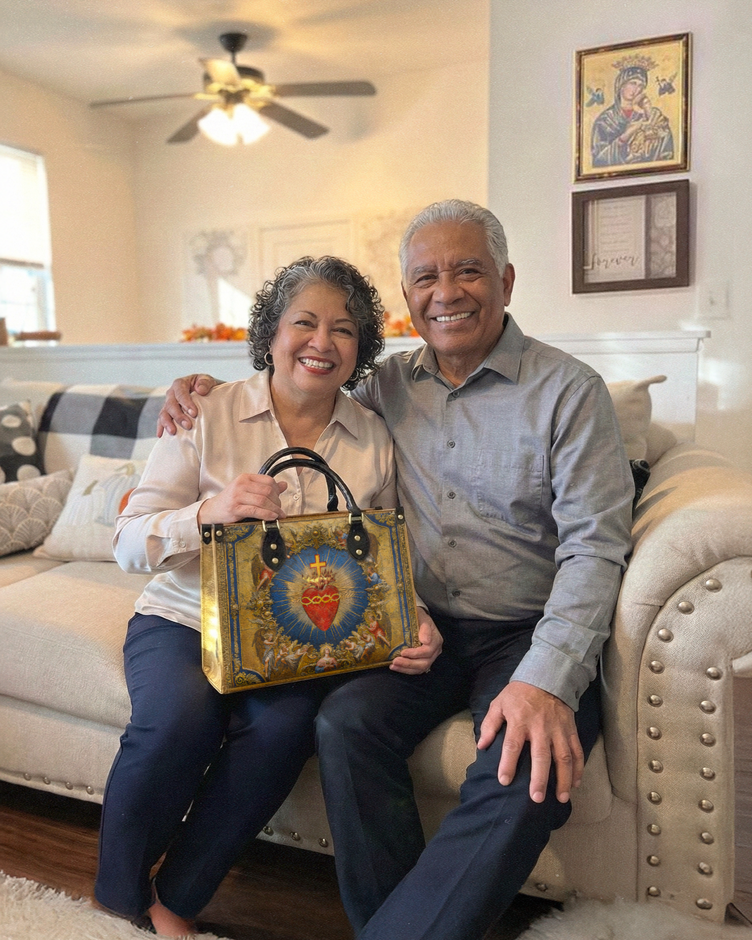 A smiling older couple sits on a couch; the woman holds a handbag with a religious image.