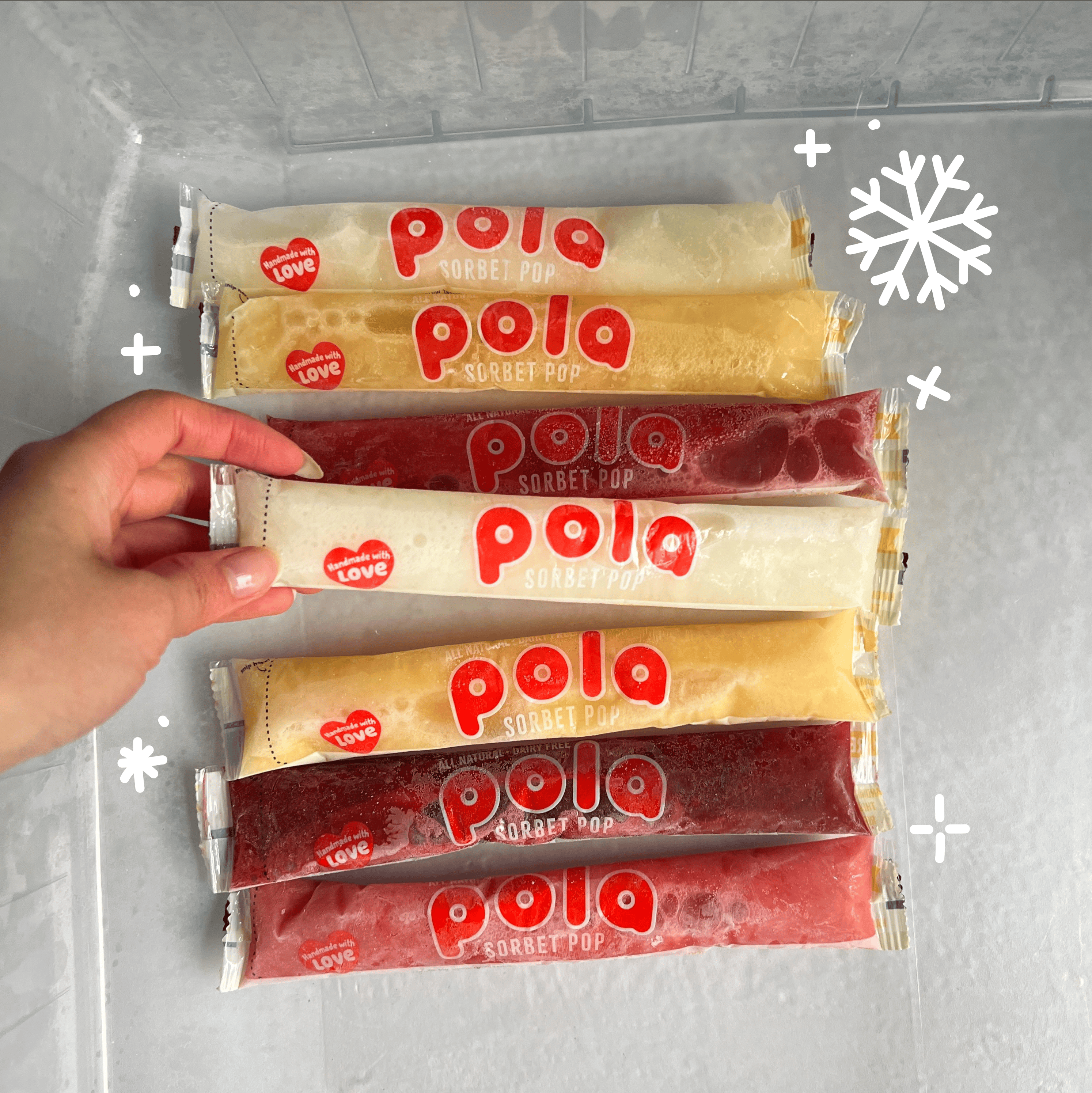 A hand selecting one of several colorful Pola Sorbet Pops from inside a freezer drawer.