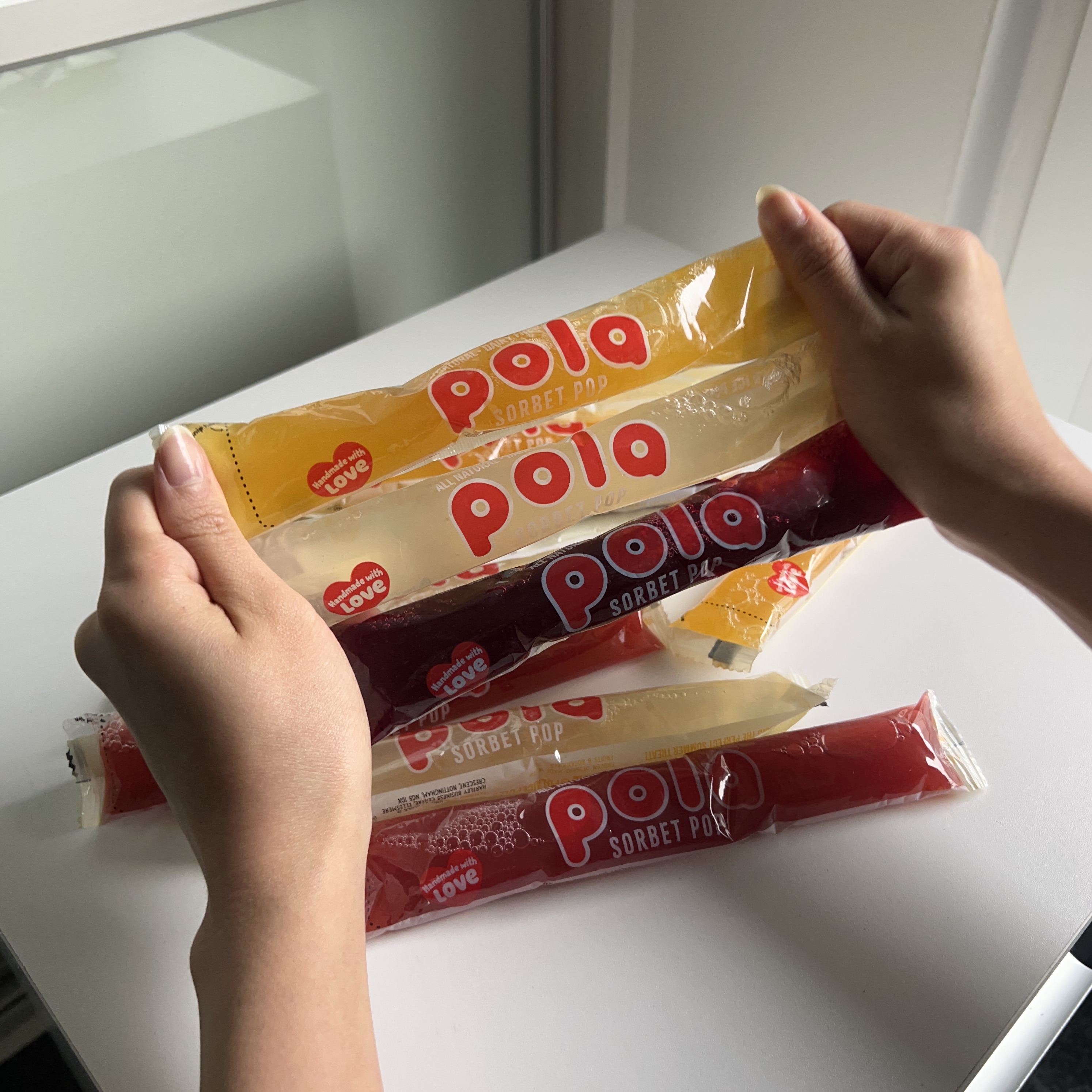 A pair of hands holding several colorful sorbet pops in plastic tubes over a white surface.