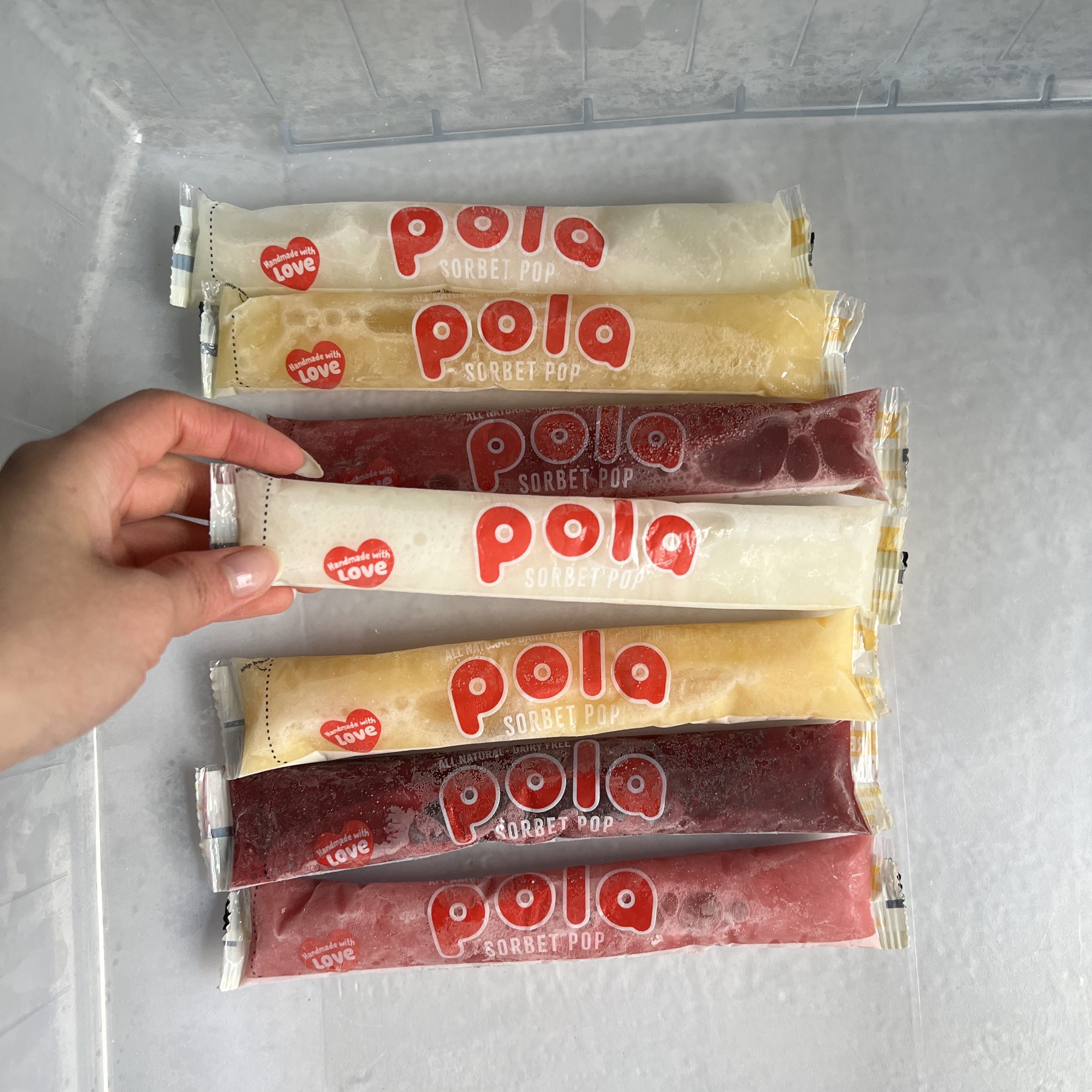A hand selecting one of several colorful Pola Sorbet Pops from inside a freezer drawer.