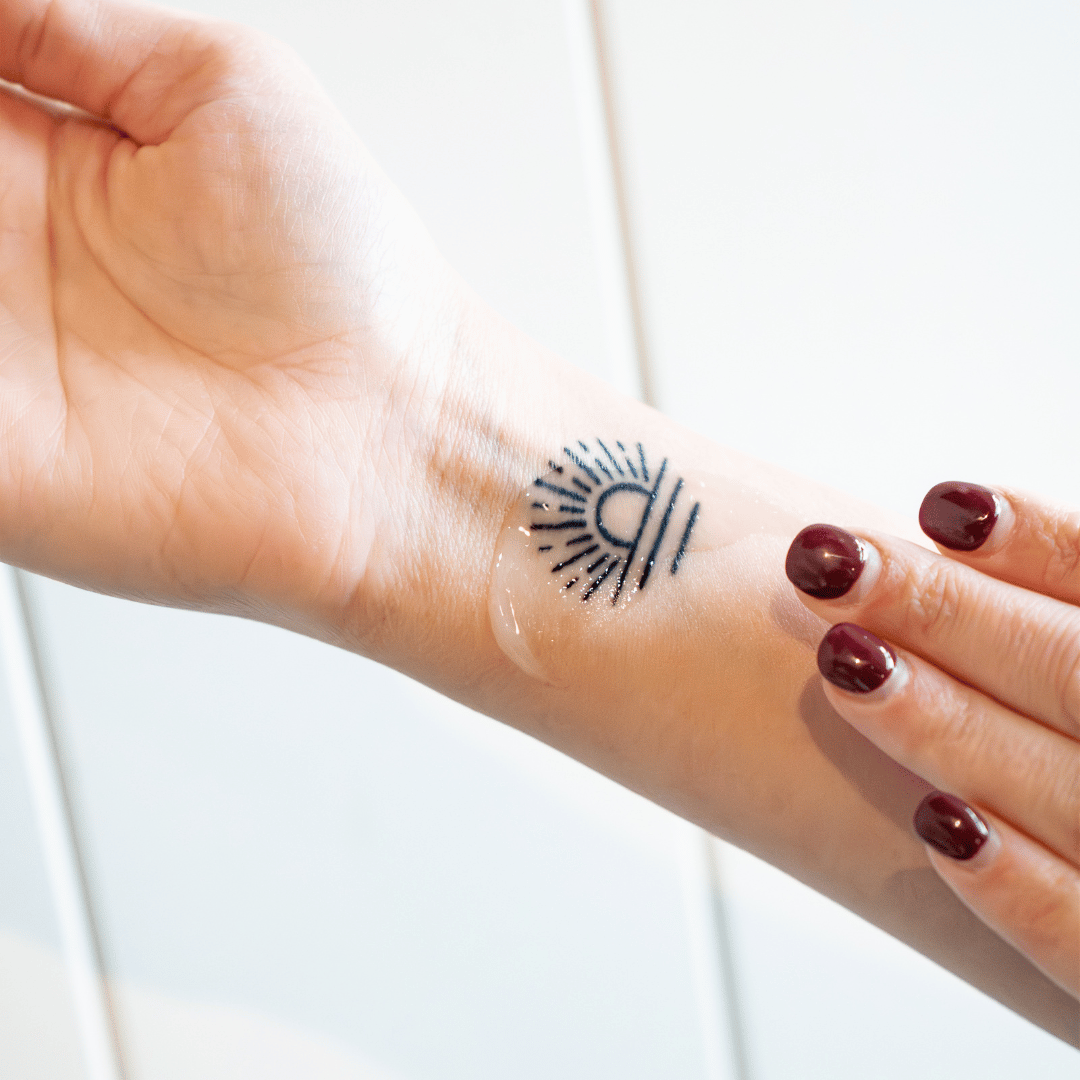 A hand with dark red nail polish applies a clear gel to a temporary sun tattoo on a wrist.