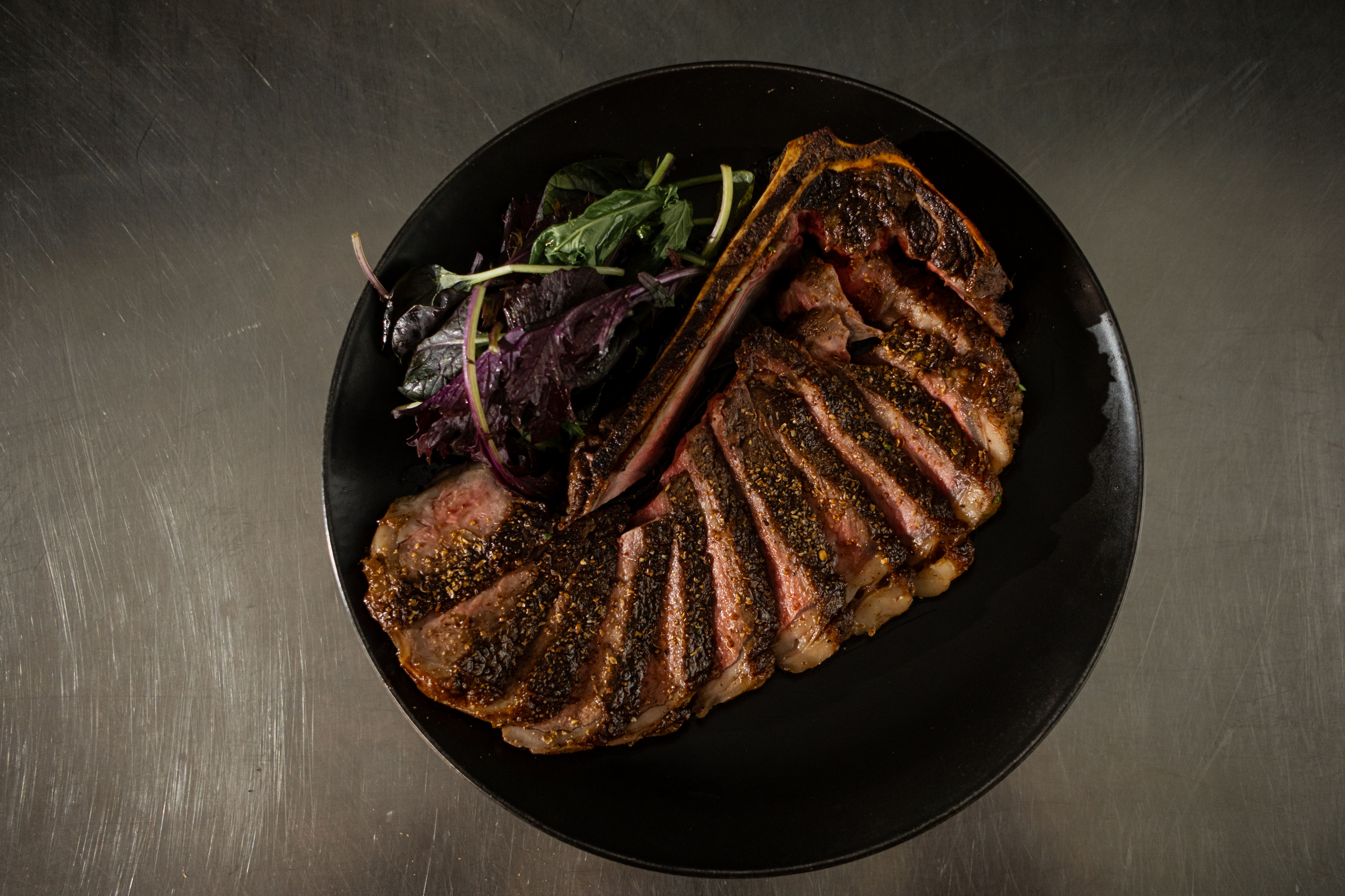 Sliced steak with seasoning and greens on a black plate.