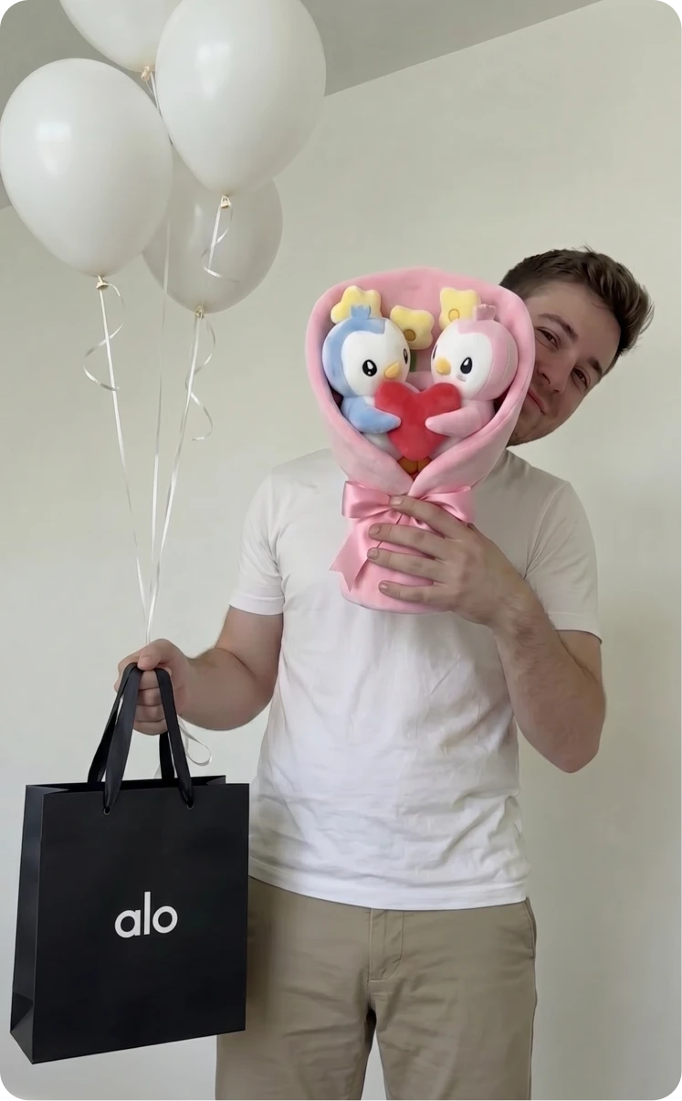 A man holds a bouquet of plush penguins, a black shopping bag, and white balloons.