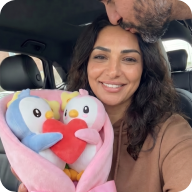 A woman in a car holds a bouquet of stuffed penguins while a man kisses her forehead.