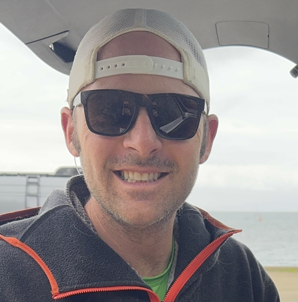 Person wearing sunglasses and a backwards cap, smiling outdoors.
