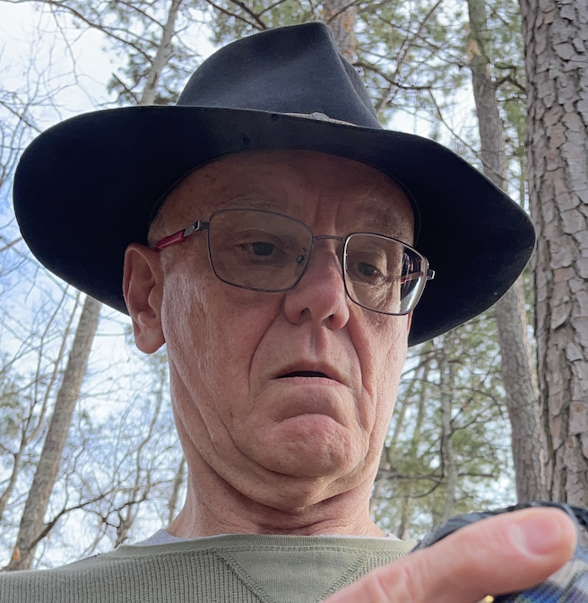 Person in a black hat and glasses, looking at a smartphone in a forest.