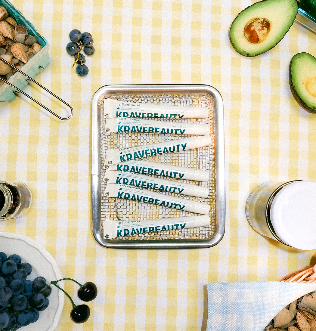 KraveBeauty Lip Barrier Relief tubes in a basket on a picnic blanket surrounded by fruit and nuts.